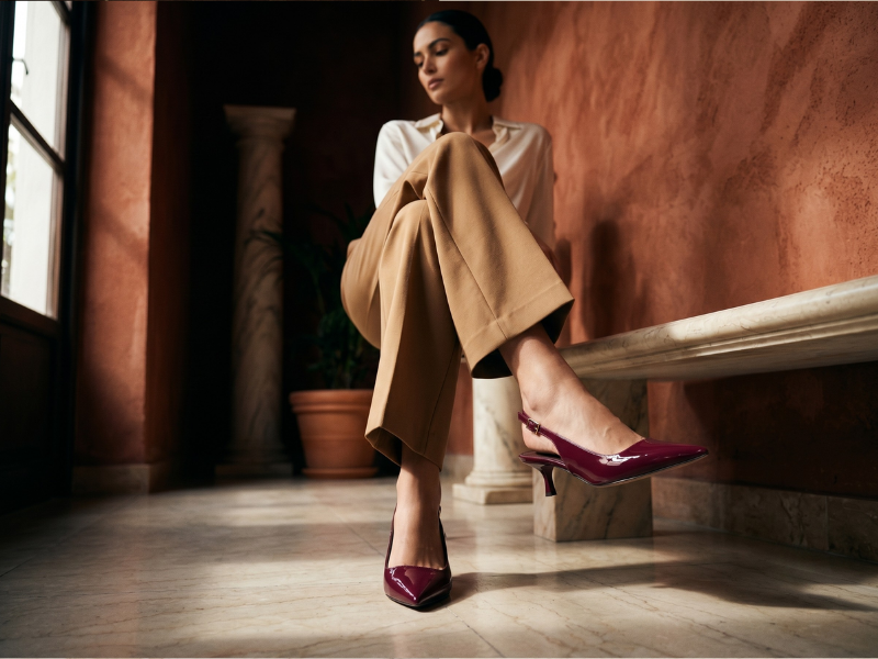 Mulher sentada em ambiente de paredes terracota usando scarpin vinho de verniz com bico fino em destaque, sob luz natural lateral
